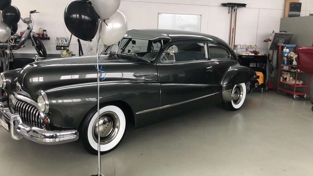 Ein langer schwarzer frisch lackierter Oldtimer. Davor sind zwei Stangen mit jeweils einigen Ballons in Silber und Schwarz.