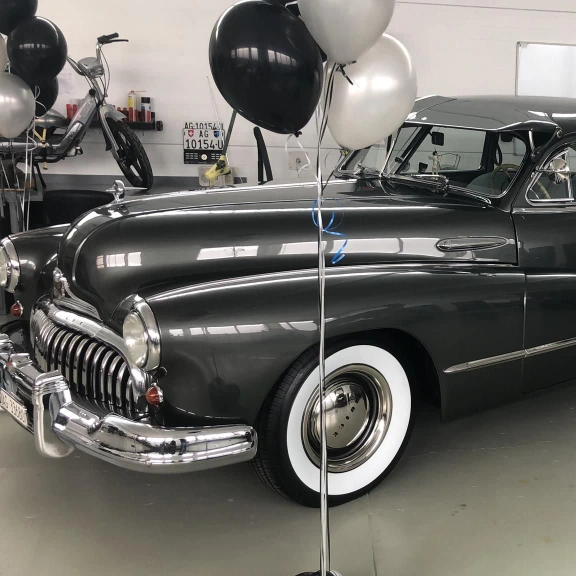 Ein langer schwarzer frisch lackierter Oldtimer. Davor sind zwei Stangen mit jeweils einigen Ballons in Silber und Schwarz.